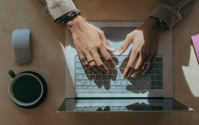 Hände tippen auf der Tastatur eines offenen Laptops auf einem Schreibtisch; daneben stehen eine Kaffeetasse, eine Computermaus und farbige Notizbücher im Tageslicht.
