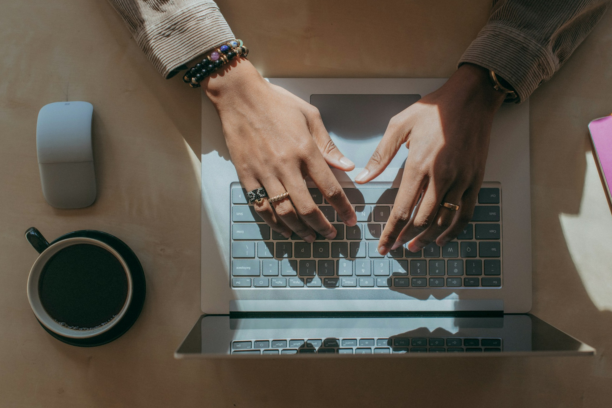 Hände tippen auf der Tastatur eines offenen Laptops auf einem Schreibtisch; daneben stehen eine Kaffeetasse, eine Computermaus und farbige Notizbücher im Tageslicht.